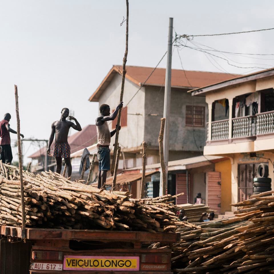 Workers loading long wooden logs onto a truck in a busy street with buildings and power lines in the background.