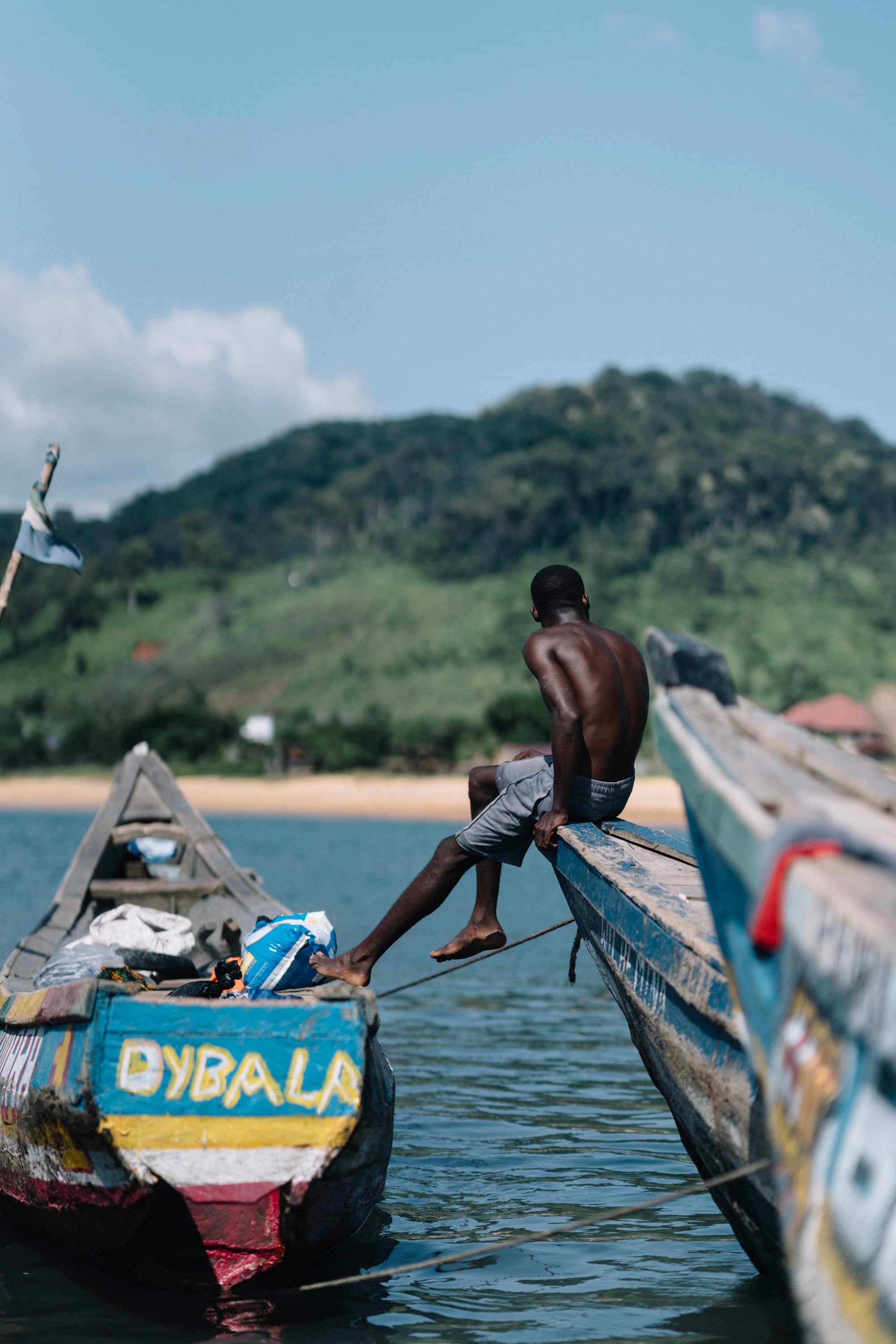 A man sits on the edge of a colorful wooden boat on a calm lake, with a lush green hill in the background under a clear blue sky.